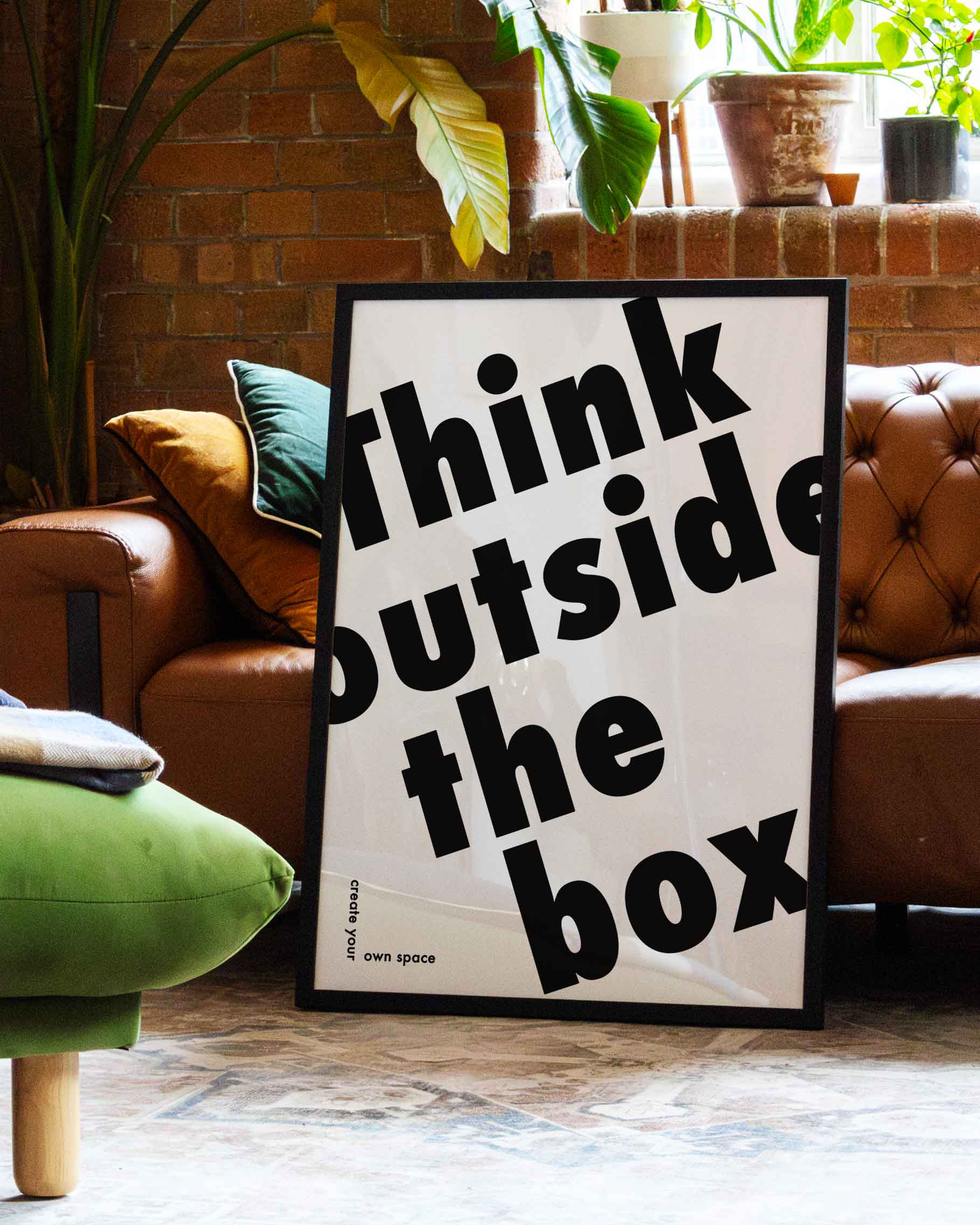 Outside the Box | Typografie Poster – Spawnd® in schwarzem Rahmen, dekorativ inszeniert in einem stilvollen Wohnzimmer mit Pflanzen und Ledersofa.